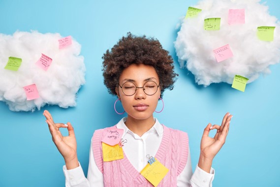 Light blue background with fake white clouds. A girl with brown skin and hair is in the center with her eyes closed and hands up in a meditative position. She is wearing round glasses, hoop earrings, a white collared shirt with a pink v neck sweater vest.