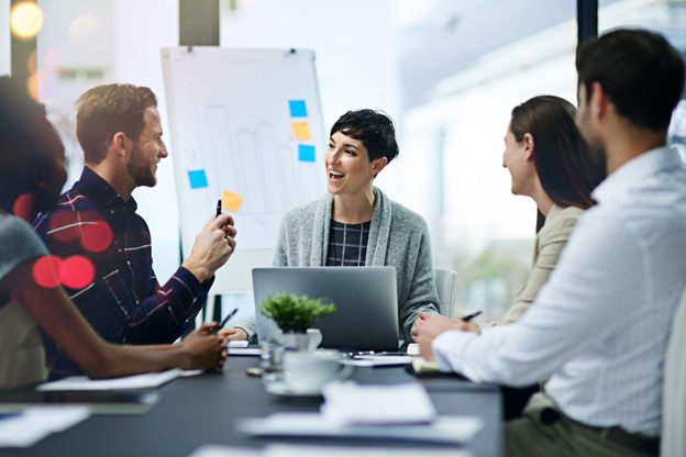 you are looking straight to a group of three people sitting at a conference table. the person in the center is looking to their right and there is a laptop infront of them.