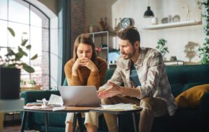 A man and woman sit on a couch, focused on a laptop screen between them.