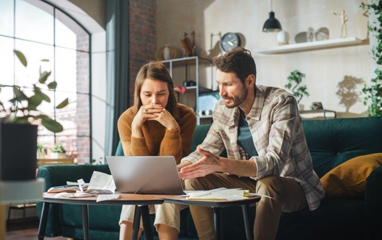 A man and woman sit on a couch, focused on a laptop screen between them.