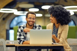 Two young happy multiracial business people sitting at the desk in the modern office, using laptop and discussing project, man and woman working together in coworking space