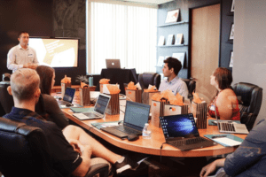 group of people sitting around an oval table with lap tops open. they are looking in the same direction where a teacher is standing