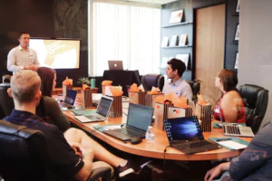 group of people sitting around an oval table with lap tops open. they are looking in the same direction where a teacher is standing