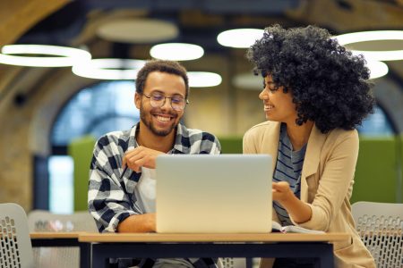 Two young happy multiracial business people sitting at the desk in the modern office, using laptop and discussing project, man and woman working together in coworking space