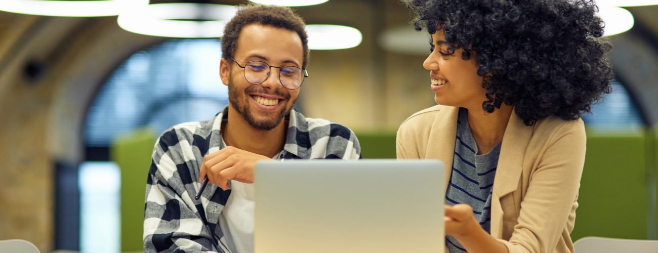 Two young happy multiracial business people sitting at the desk in the modern office, using laptop and discussing project, man and woman working together in coworking space