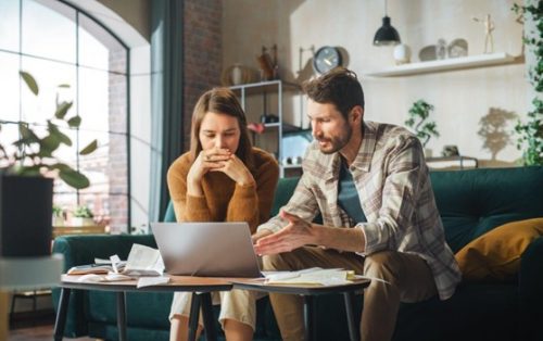 A man and woman sit on a couch, focused on a laptop screen between them.
