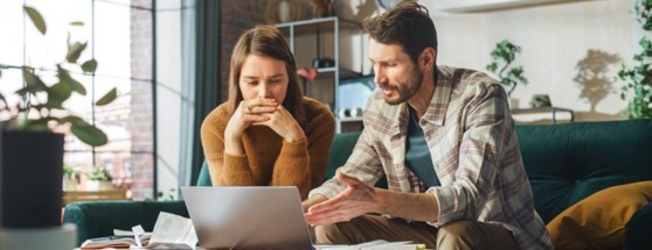 A man and woman sit on a couch, focused on a laptop screen between them.