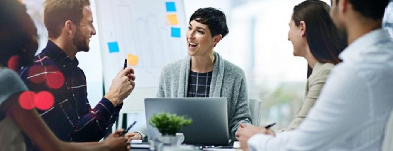 you are looking straight to a group of three people sitting at a conference table. the person in the center is looking to their right and there is a laptop infront of them.