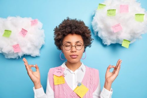 Light blue background with fake white clouds. A girl with brown skin and hair is in the center with her eyes closed and hands up in a meditative position. She is wearing round glasses, hoop earrings, a white collared shirt with a pink v neck sweater vest.