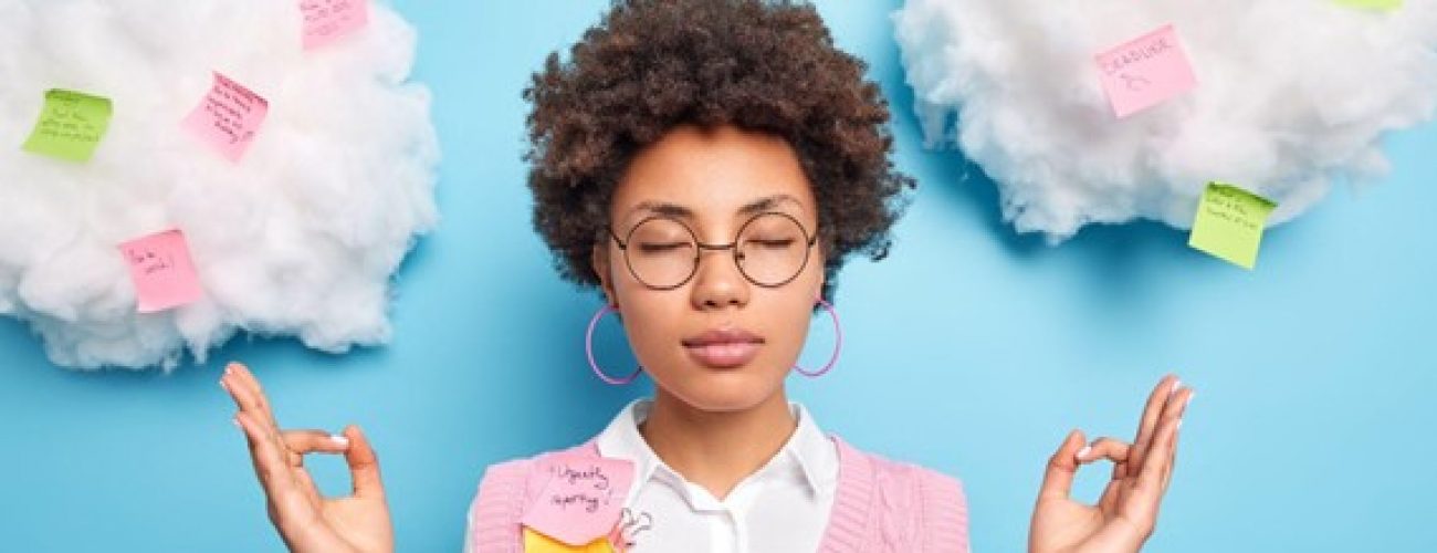 Light blue background with fake white clouds. A girl with brown skin and hair is in the center with her eyes closed and hands up in a meditative position. She is wearing round glasses, hoop earrings, a white collared shirt with a pink v neck sweater vest.