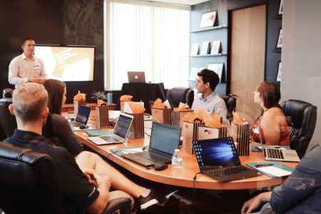 group of people sitting around an oval table with lap tops open. they are looking in the same direction where a teacher is standing