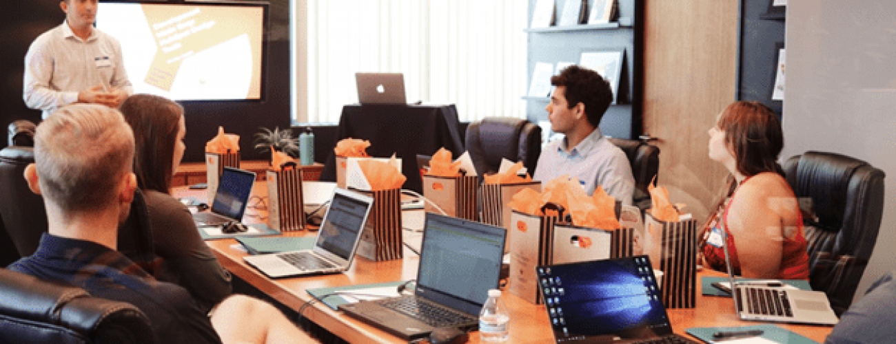 group of people sitting around an oval table with lap tops open. they are looking in the same direction where a teacher is standing